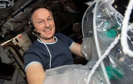 Ein Astronaut mit Headset lächelt im Inneren eines Raumfahrzeugs neben einer transparenten Verpackung mit grünem Inhalt.