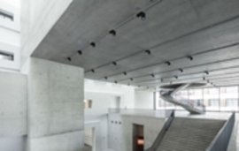 Modernes Gebäudeinnere mit massiven Betonwänden, einer schrägen Treppe und einem hohen Deckengewölbe aus Sichtbeton.