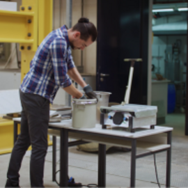 Ein Mann in einem karierten Hemd arbeitet mit Handschuhen an einem Behälter auf einem Tisch im Labor, neben ihm steht eine technische Vorrichtung.