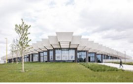 Modernes Gebäude mit markanter, gestaffelter Dachkonstruktion und großer Glasfassade, umgeben von einer grünen Wiese unter einem bewölkten Himmel.