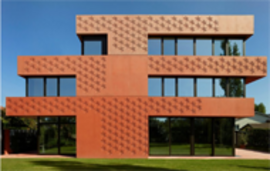 Ein modernes mehrstöckiges Gebäude mit einer texturierten, rötlich-orangen Fassade, großen Glasfenstern und einem grünen Rasen davor vor blauem Himmel.