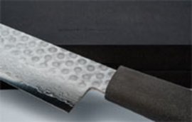 Ein handgeschmiedetes Kochmesser mit geädertem Metallblatt und dunklem Griff liegt schräg auf einer hellen Oberfläche vor schwarzem Hintergrund.