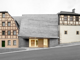 Modernes Betonhaus mit großer Dachfläche flankiert von traditionellen Fachwerkhäusern